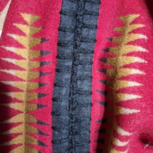 Puede incluir: Primer plano de un textil rojo con un patrón geométrico en dorado y negro. Se ve una costura vertical negra central, que añade contraste de textura. La tela parece gruesa y tejida, posiblemente lana.