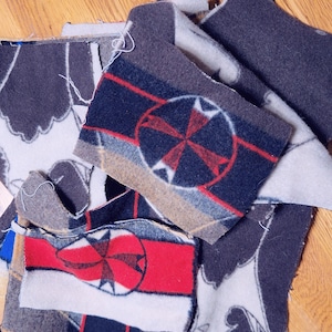 May include: Close-up of several fabric patches with geometric designs. The patches feature a color palette of red, navy blue, gray, and white. Some patches have a compass rose design. The patches are on a wooden surface.