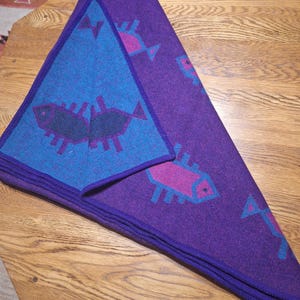 May include: A folded purple blanket with a blue triangle on top. The blanket features a repeating pattern of stylized fish in pink and blue. The blanket is folded on a wooden surface.