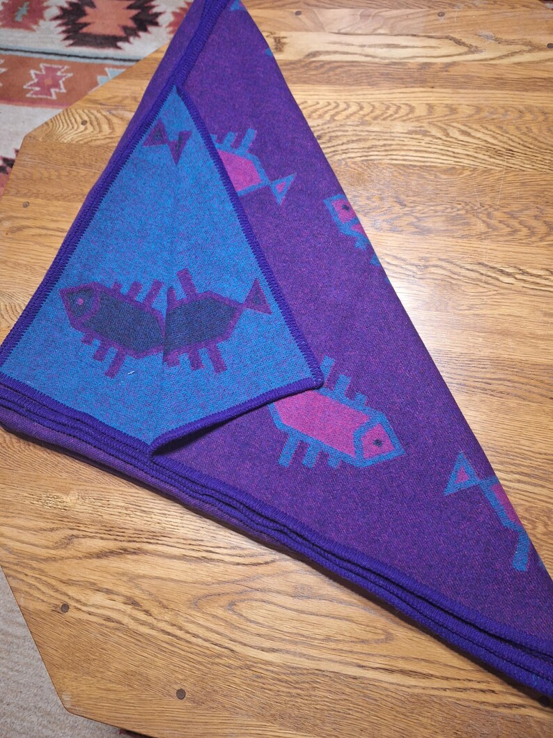May include: A folded purple blanket with a blue triangle on top. The blanket features a repeating pattern of stylized fish in pink and blue. The blanket is folded on a wooden surface.