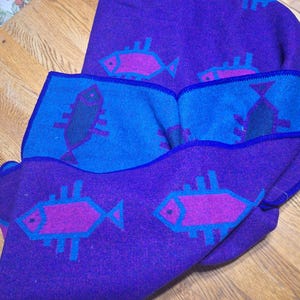 May include: A folded purple blanket with a blue border and fish pattern. The fish are pink and outlined in blue. The blanket is made of a soft material and is folded over on a wooden surface.