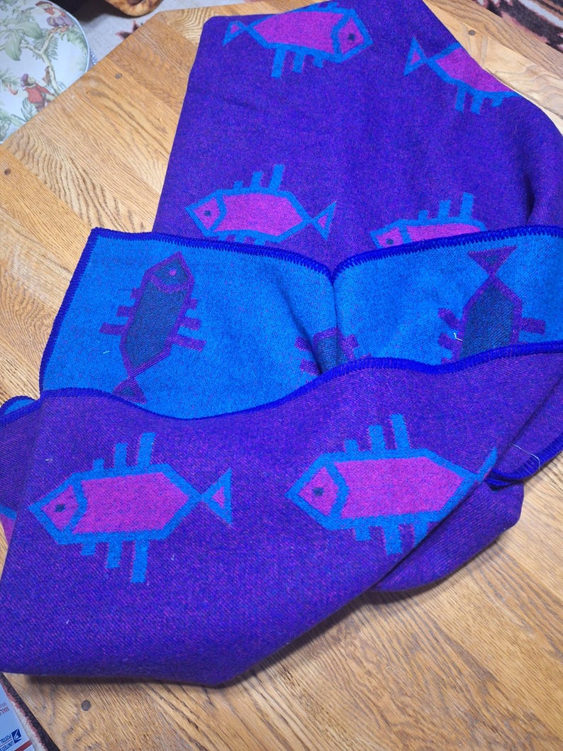 May include: A folded purple blanket with a blue border and fish pattern. The fish are pink and outlined in blue. The blanket is made of a soft material and is folded over on a wooden surface.