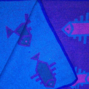 May include: A folded textile with a fish pattern. The fabric is predominantly blue and purple, with a dark blue border. The fish are depicted in a stylized, geometric design, with purple and green accents. The textile is folded to reveal both sides.