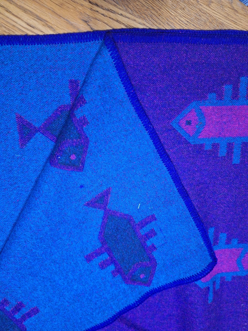 May include: A folded textile with a fish pattern. The fabric is predominantly blue and purple, with a dark blue border. The fish are depicted in a stylized, geometric design, with purple and green accents. The textile is folded to reveal both sides.