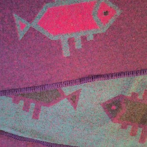 May include: A close-up of a purple textile with a repeating fish pattern. The fish are outlined in teal and filled with magenta. The textile has a textured appearance and a stitched border.
