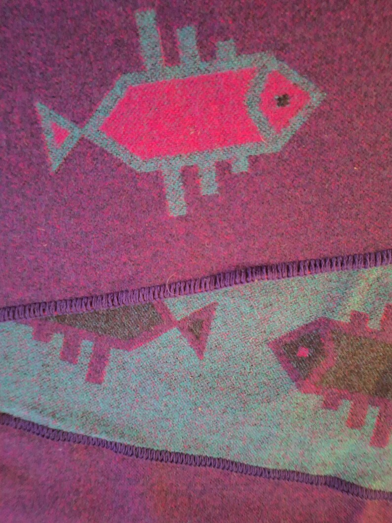 May include: A close-up of a purple textile with a repeating fish pattern. The fish are outlined in teal and filled with magenta. The textile has a textured appearance and a stitched border.