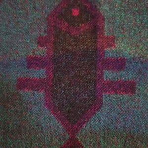 May include: Textured fabric in teal and green, featuring a central geometric design in dark purple. The design, resembling a fish, has a dark core and a lighter purple outline. The fabric's texture is visible.