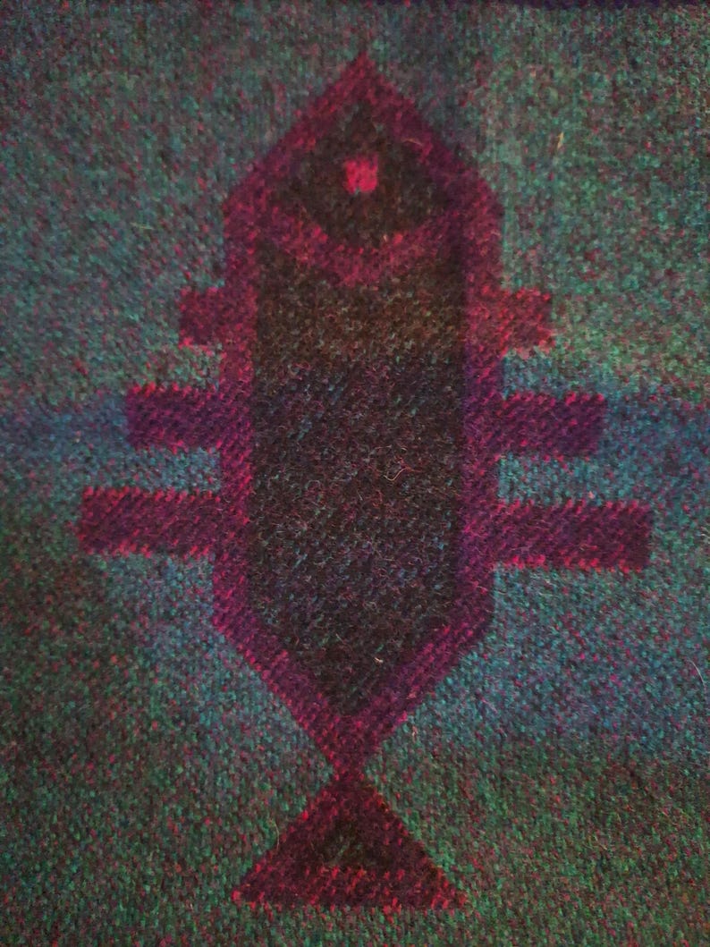 May include: Textured fabric in teal and green, featuring a central geometric design in dark purple. The design, resembling a fish, has a dark core and a lighter purple outline. The fabric's texture is visible.