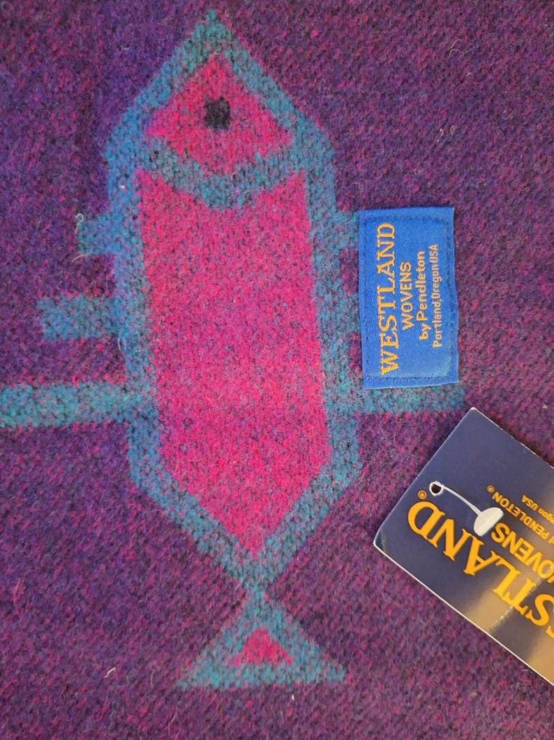 May include: Close-up of a woven textile with a geometric design in shades of purple, pink, and teal. The design features a central arrow-like shape. A blue tag reads "WESTLAND WOVENS by Pendleton Portland, Oregon USA."