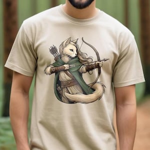 Könnte beinhalten: Ein beiges T-Shirt mit einer weißen Katze, die einen grünen Umhang mit Kapuze trägt und einen Bogen und Pfeil hält. Die Katze zielt mit dem Pfeil und hat einen ernsten Gesichtsausdruck.