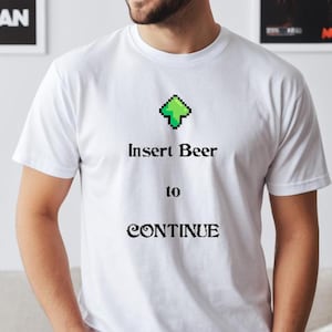 Könnte beinhalten: Weißes T-Shirt mit einem grünen, pixelartigen Pfeil, der nach oben zeigt, und dem Text "Insert Beer to CONTINUE", der darauf gedruckt ist.
