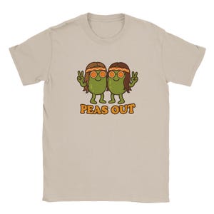 Könnte beinhalten: Beiges T-Shirt mit einem Cartoon-Design von zwei grünen Erbsen mit braunen Haaren, orangefarbenen Sonnenbrillen und Friedenszeichen. Der orangefarbene Text "PEAS OUT" ist darunter gedruckt. Das T-Shirt hat einen Rundhalsausschnitt und kurze Ärmel.