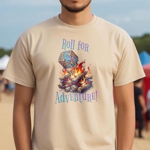 Könnte beinhalten: Ein beiges T-Shirt mit einem Aufdruck eines mehrfarbigen zwanzigseitigen Würfels auf einem Lagerfeuer. Der Text "Roll for Adventure!" ist unter dem Aufdruck gedruckt.