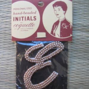 May include: A pink beaded letter E, with the text "Personalized hand-beaded INITIALS coquette of Hollywood, California" on a white and red card with a woman's illustration.