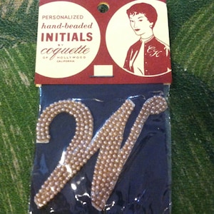 May include: A white, beaded letter W, in a cursive font, is packaged in a clear plastic bag. The bag is attached to a red and white card that reads "Personalized hand-beaded INITIALS by coquette of HOLLYWOOD CALIFORNIA". A woman with dark hair is pictured on the card, wearing a blue jacket with the initials "C.H." embroidered on the front.