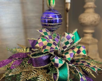 May include: A decorative centerpiece featuring a purple and green ornament with a gold base. The ornament has a purple glass ball with gold stripes, topped with a green and gold finial. The base is adorned with a large bow and glittery foliage.