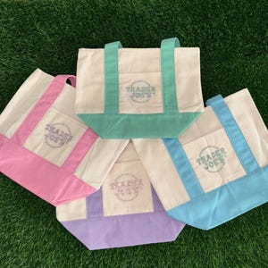 May include: A collection of canvas tote bags in various pastel colors: pink, light blue, mint green, and lavender. Each bag features a front pocket and handles in a matching color, with the "Trader Joe's" logo printed on the front.