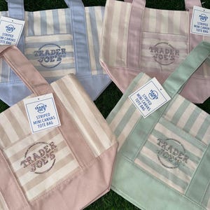 May include: Four mini canvas tote bags in light blue, pink, tan, and green, each featuring vertical stripes, a front pocket, and the text Trader Joe's embroidered in glittery thread. Each bag includes a white tag labeled Striped Mini Canvas Tote Bag.