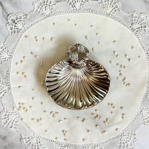 Puede incluir: Un plato plateado en forma de concha con un diseño acanalado, sobre un mantel individual blanco con bordes de encaje. Pequeñas cuentas con forma de perla están dispersas alrededor del plato. El fondo es una superficie de mármol.