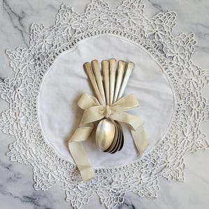 Könnte beinhalten: Ein Set aus fünf silberfarbenen Metalllöffeln, die fächerförmig angeordnet und mit einem cremefarbenen Stoffband zusammengebunden sind. Die Löffel haben glatte, konische Griffe und ovale Laffen und liegen auf einer runden, weißen Stoffdeckchen.