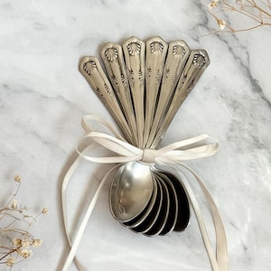 Puede incluir: Un conjunto de cucharas de plata antiguas, dispuestas en forma de abanico y atadas con una cinta color crema. Las cucharas tienen diseños grabados en los mangos. La disposición se presenta sobre un fondo de mármol.