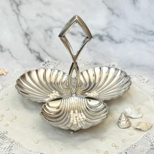 May include: A silver-colored, three-tiered serving dish with a handle. The dish has a seashell design and is filled with small, white pearls. The dish is on a white, lace-edged doily.