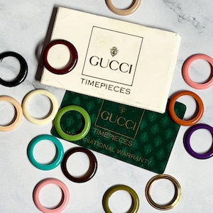 Può includere: Una collezione di quindici anelli circolari in vari colori solidi tra cui arancione, beige, nero, bordeaux, rosa, crema, verde, ottanio, marrone, viola e oro, disposti attorno a una scatola Gucci Timepieces e una carta di garanzia verde.