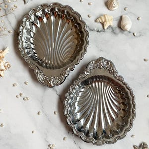 May include: Two silver shell-shaped trays with a scalloped edge and decorative detailing. The trays have a reflective surface and are set on a marble-patterned surface, surrounded by small pearls and seashells. A decorative item for the home.