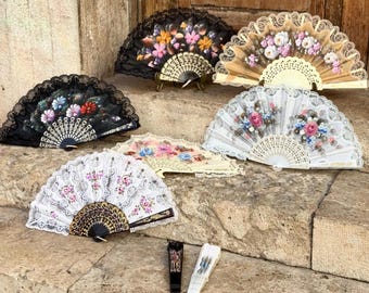 May include: A collection of seven folding hand fans displayed in various states of openness. The fans feature intricate lace detailing, floral patterns in shades of pink, blue, and yellow, and decorative plastic or wooden frames in black, white, and beige.