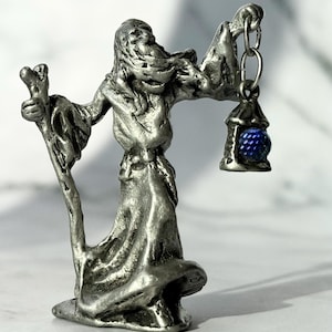 May include: A silver-colored figurine of a robed wizard holding a staff and a lantern. The lantern has a blue gem. The wizard's robe is flowing, and the figure has a detailed face and hair. The figurine is on a white surface.