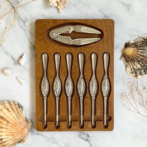 May include: A wooden serving tray with a silver crab cracker and six silver seafood forks. The forks and cracker are nestled in a wooden tray. The tray is surrounded by seashells and dried plants on a marble surface.