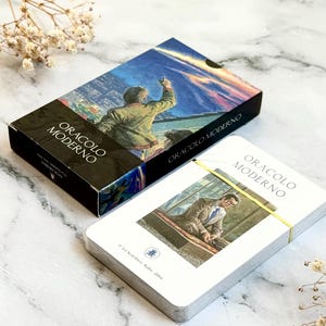 May include: A deck of "Oracolo Moderno" tarot cards with a box featuring a painting of a person pointing upwards. The card deck is bound with a yellow band. The box and cards are on a marble surface.