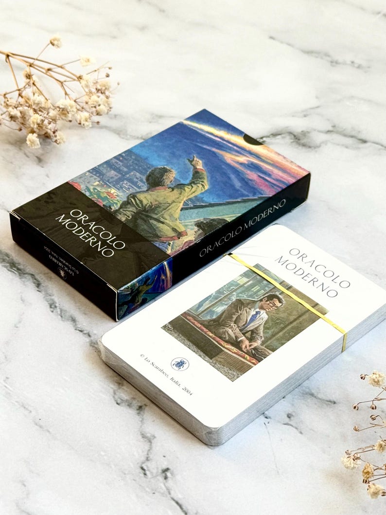 May include: A deck of "Oracolo Moderno" tarot cards with a box featuring a painting of a person pointing upwards. The card deck is bound with a yellow band. The box and cards are on a marble surface.