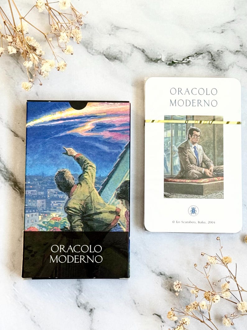 May include: A deck of oracle cards and a single card. The card box features a painting of a person pointing at a meteor shower over a city. The text "ORACLO MODERNO" is at the bottom. The single card has the same text.