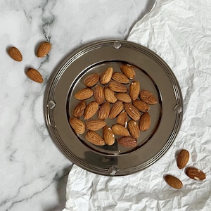 Peut inclure: Une assiette argentée remplie d'amandes crues, avec quelques-unes éparpillées. L'assiette a un bord décoratif et repose sur une surface blanche texturée. Les amandes sont de couleur brun clair.