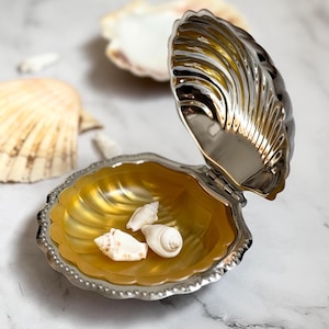 Könnte beinhalten: Eine silberne Muschel-Schmuckschatulle mit gelbem Innenraum. Die Schachtel ist geöffnet und enthält drei kleine weiße Muscheln.