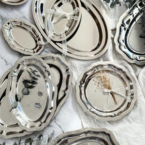 Puede incluir: Una variedad de bandejas plateadas de diferentes formas y tamaños. Algunas son ovaladas, otras redondas con bordes festoneados. Varias están atadas con cinta de encaje blanco. Ramas de eucalipto añaden un toque decorativo.