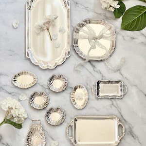 Puede incluir: Una variedad de bandejas y platos de servir plateados ornamentados en varias formas y tamaños. La colección incluye piezas rectangulares, ovaladas y en forma de concha, algunas con asas y bordes decorativos. Flores y pétalos blancos están esparcidos alrededor de las bandejas.