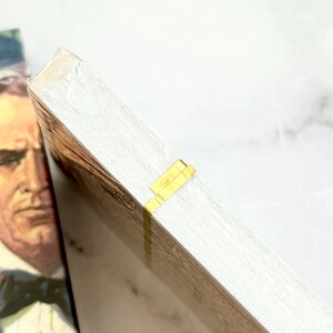 May include: A stack of white paper with a gold-colored tape closure. The paper has a light wood grain pattern. A portion of a book cover with a man in a suit is visible in the background.