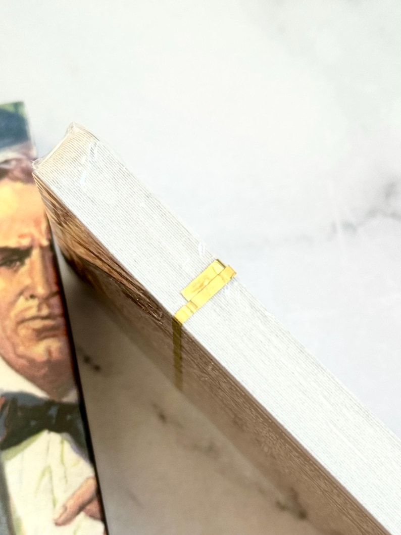 May include: A stack of white paper with a gold-colored tape closure. The paper has a light wood grain pattern. A portion of a book cover with a man in a suit is visible in the background.
