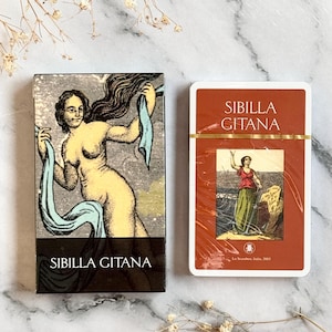 May include: Two decks of Sibilla Gitana tarot cards. One deck has a black background with a nude woman holding blue fabric. The other deck has a red background with a woman in a red top and green skirt. Both decks have the text "SIBILLA GITANA".