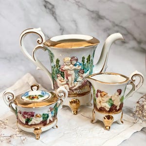 Puede incluir: Un juego de té de porcelana blanca con ribetes dorados, que incluye una tetera, azucarero y jarrita para la leche. Cada pieza está decorada con un relieve colorido de querubines y follaje. El juego se exhibe sobre un paño blanco bordado.