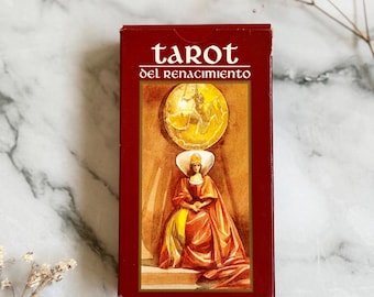 May include: A red tarot card box with the words "tarot del Renacimiento" in white. The box features an illustration of a woman in a Renaissance-style gown and a gold medallion. The box is rectangular and sits on a marble surface.