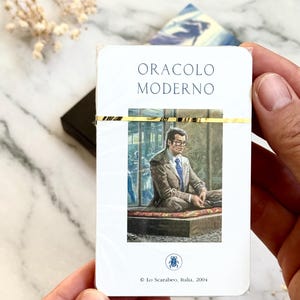May include: A deck of cards titled "ORACLO MODERNO" with a gold band. The card illustration depicts a man in a suit. The deck is held by hands against a marble background.