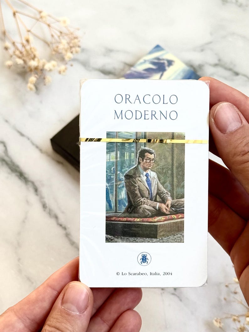 May include: A deck of cards titled "ORACLO MODERNO" with a gold band. The card illustration depicts a man in a suit. The deck is held by hands against a marble background.
