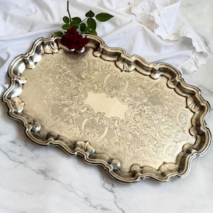 Op de afbeelding: Een zilverkleurige, sierlijke serveerschaal met een decoratieve, geschulpte rand. De schaal heeft een gegraveerd bloemmotief en een rechthoekige vorm. Een rode roos is op de schaal geplaatst. De schaal ligt op een witte doek.