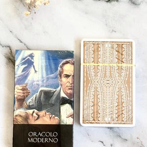 May include: A tarot card deck and box set. The box features a vintage illustration of a man and woman with the text "ORACLO MODERNO". The card backs have a beige background with a white decorative pattern. A yellow band wraps the cards.