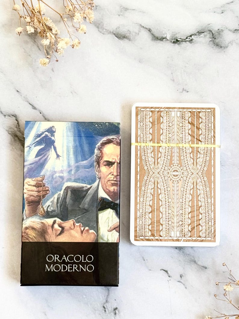 May include: A tarot card deck and box set. The box features a vintage illustration of a man and woman with the text "ORACLO MODERNO". The card backs have a beige background with a white decorative pattern. A yellow band wraps the cards.