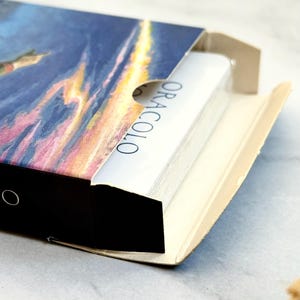 May include: A deck of cards in a box with a colorful illustration of a sunset and a flying figure. The cards are white with the word "ORACOLO" printed on the side. The box is black and beige.