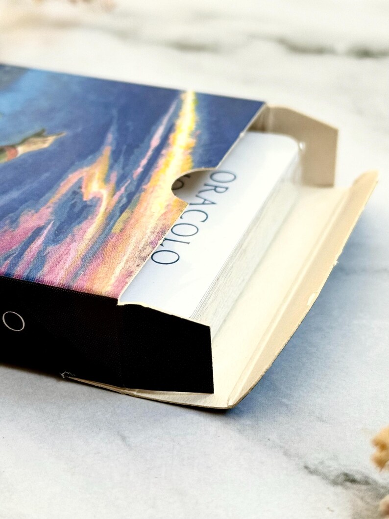 May include: A deck of cards in a box with a colorful illustration of a sunset and a flying figure. The cards are white with the word "ORACOLO" printed on the side. The box is black and beige.